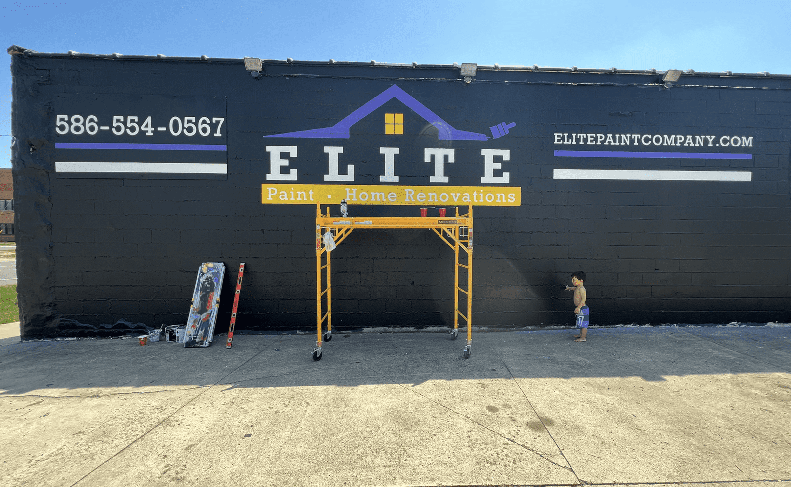 Sterling Heights, MI 10 Painted wall with Elite Paint and Home Renovations logo, scaffold, tools, and child nearby.
